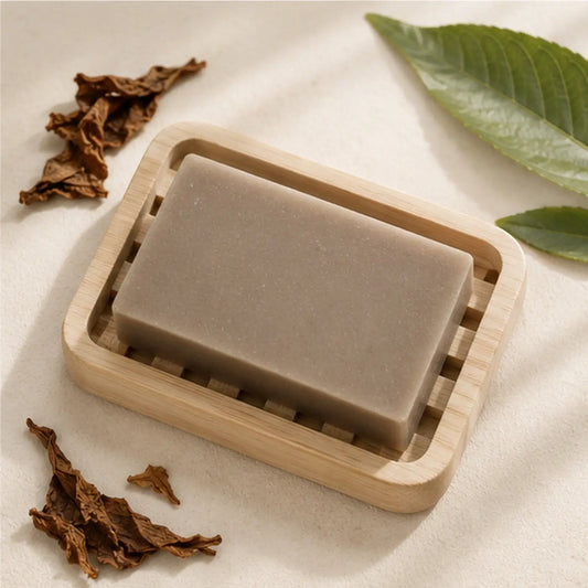 A rectangular matte gray tobacco leaf soap bar made with palm oil rests in a light wood-grain soap dish handcrafted skincare