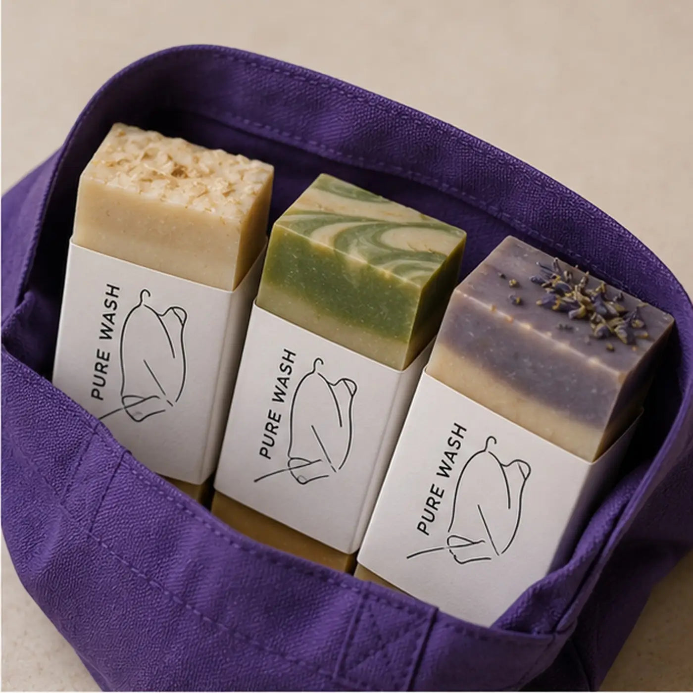 Three handcrafted soap bars in white paper sleeves inside a purple reusable tote bag soap bundle for personal care