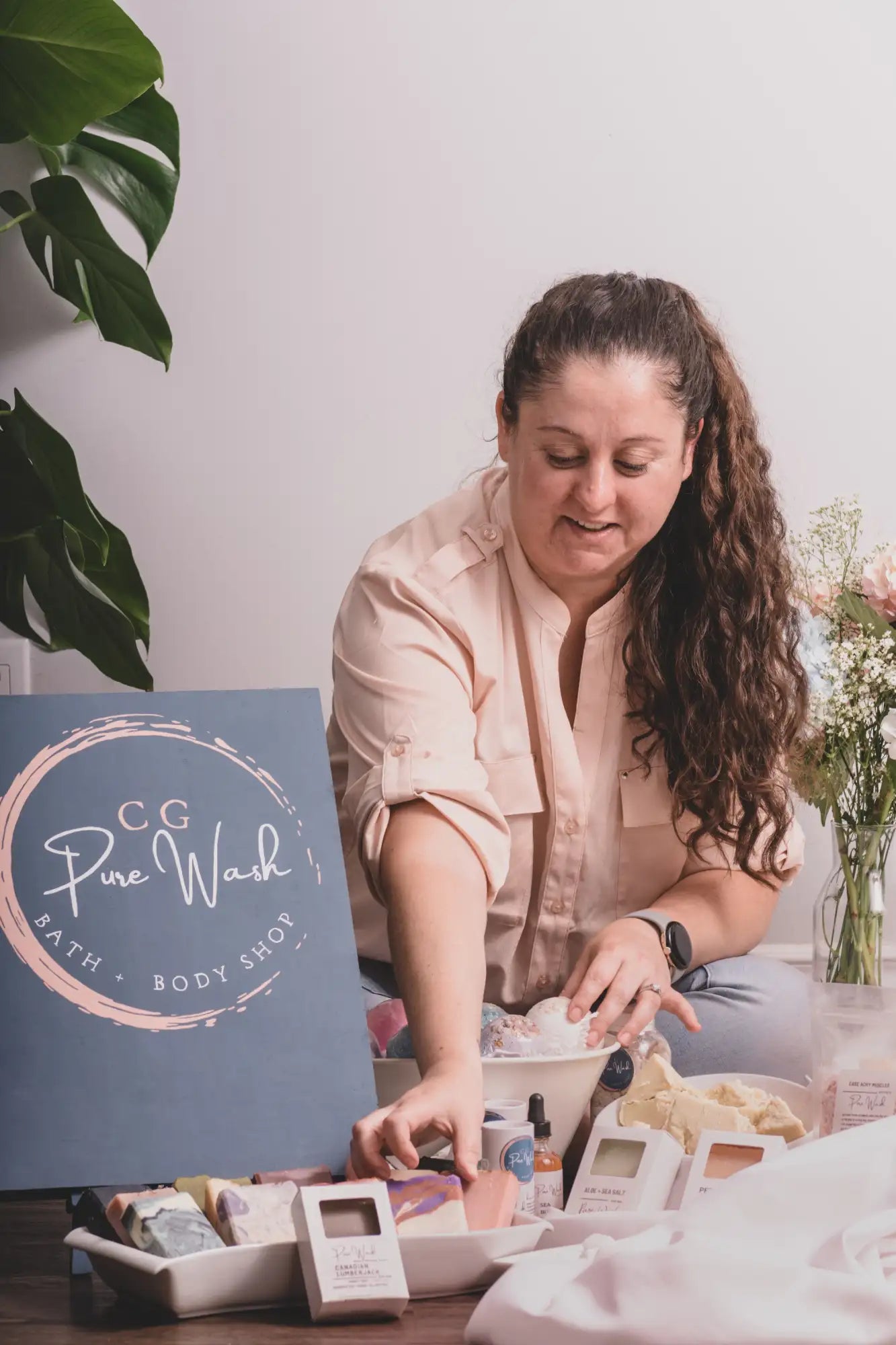 A woman in a soft pink button-up shirt sits beside a blue sign for ’cg pure wash bath + body shop,’ surrounded by an array of bath and body products including soaps, lotions, and candles.